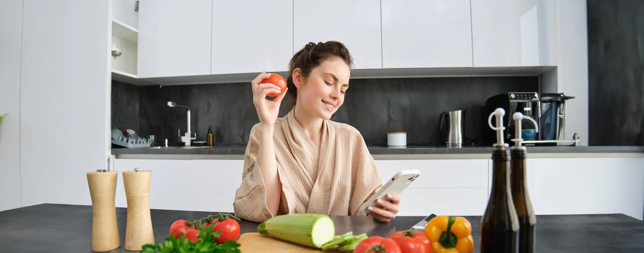 A woman in her kitchen holding a tomato and checking her phone, with fresh vegetables on the counter